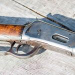 Nahaufnahme eines klassischen Unterhebelrepetierers mit Holzschaft und Metallverschluss, liegend auf verwittertem Holztisch – gut erkennbar ist der charakteristische Unterhebelmechanismus der Waffe.